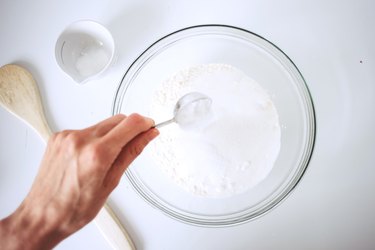 Hand spooning dry ingredients into a glass bowl.