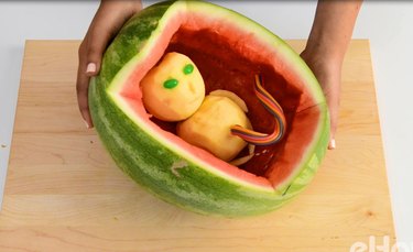 Hands holding hollowed-out watermelon with alien apple baby inside.