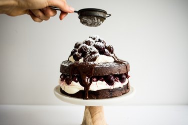 Hand-held strainer shaking powdered sugar over the chocolate ganache on the cake.