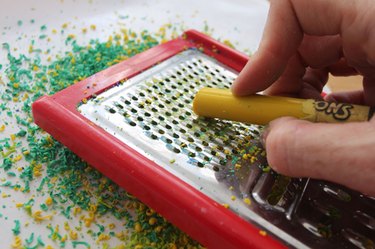 A cheese grater held over crayon shavings with a peeled crayon on top of the grater