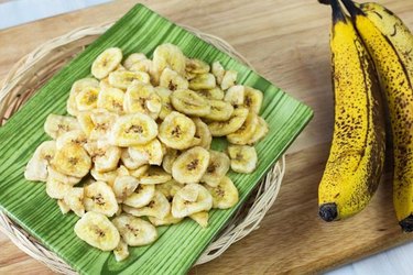 A pile of homemade banana chips next to two ripe bananas.