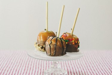 Three decorated caramel apples on a glass cake stand