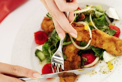 Close-up of a woman's hands slicing a piece of fried fish with fork and knife