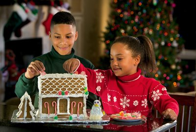 A brother and sister decorate a gingerbread house at Christmastime.