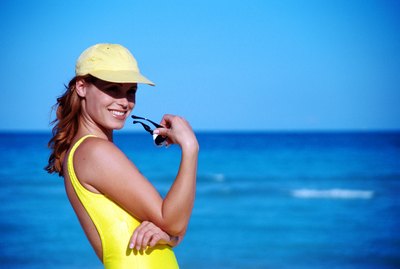 Young woman wearing cap and holding sunglasses on beach, smiling