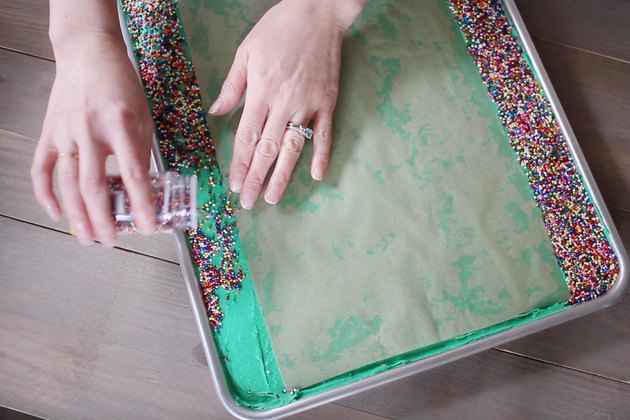 Pouring sprinkles onto sides of sheet cake while parchment paper protects center of cake