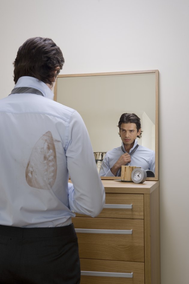 Young man adjusting tie in mirror, iron stain on shirt, rear view