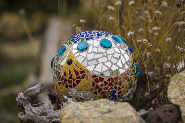 Bowling ball mortared with ceramic tiles and broken mirrors in outdoor garden. Turquoise glass buttons in sea of mirrors. Deep blue ceramic chips on top. Yellow and red ceramic chips create pattern on lower front side. Driftwood chunk lower left and dried flowers behind right.