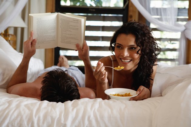 Woman with cereal bowl in bed, man reading, smiling, close-up