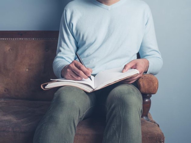 A young man is sitting on an old sofa and is writing in a notebook