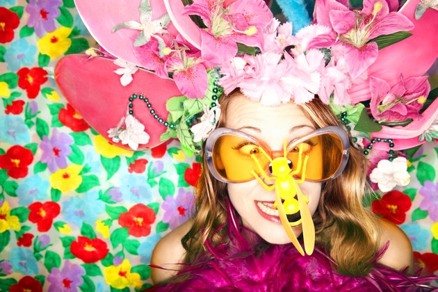 A woman looks cross-eyed at the plastic bug on her nose while wearing a wacky hat of flowers and orange sunglasses in front of a flowered background.