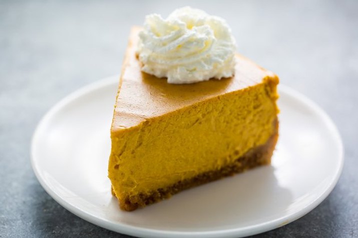 A thick wedge of pumpkin cheesecake with a whipped cream rosette on top, served on a white plate and a light-grey counter