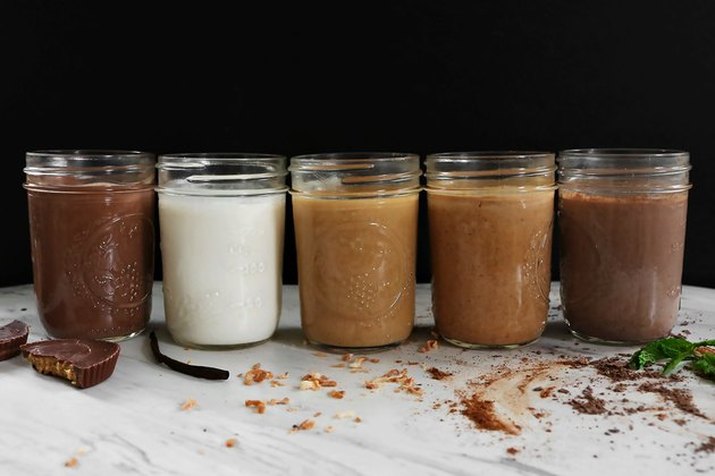 A row of homemade coffee creamers in different flavors, served in mason jars, and with flavoring ingredients scattered around them on a white tablecloth