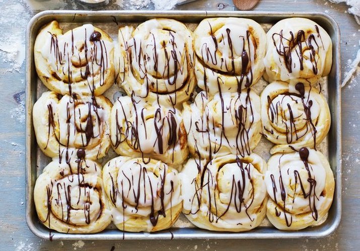 An overhead shot of a dozen pumpkin cinnamon rolls on a baking sheet, drizzled with icing and chocolate