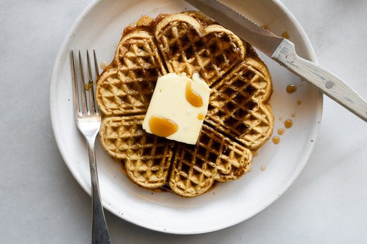 Waffles made in a decorative iron, shot from overhead, on a white plate with a pat of butter, syrup, knife and fork