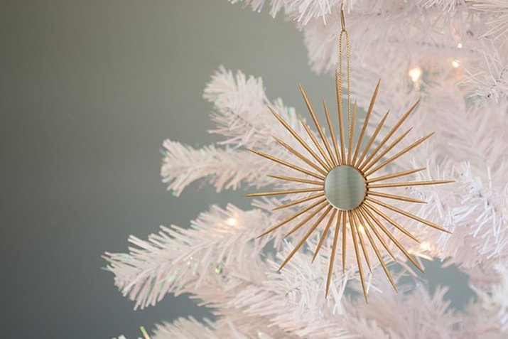DIY ornament consisting of a small circular mirror surrounded in a sunburst pattern by gold-painted toothpicks, hanging from a white Christmas tree