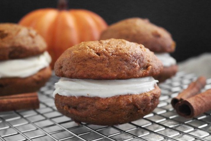 Pumpkin-spiced whoopie pies on a wire cooling rack, with cinnamon sticks visible in the foreground and a blurred pumpkin in the background