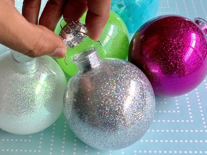 A cluster of four handmade glitter ornaments on a blue tablecloth, with a hand performing final assembly on the foremost ornament