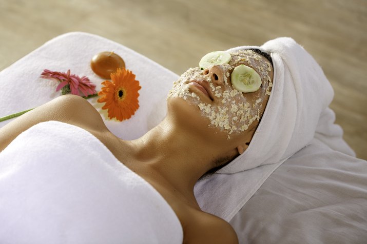 Hispanic young woman laying on bed and getting oatmeal face scrub and resting with cucumber slices on eyes.