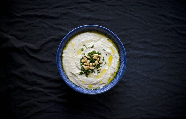 A bowl of creamy white bean hummus topped with a drizzle of olive oil, toasted pine nuts and chopped parsley.
