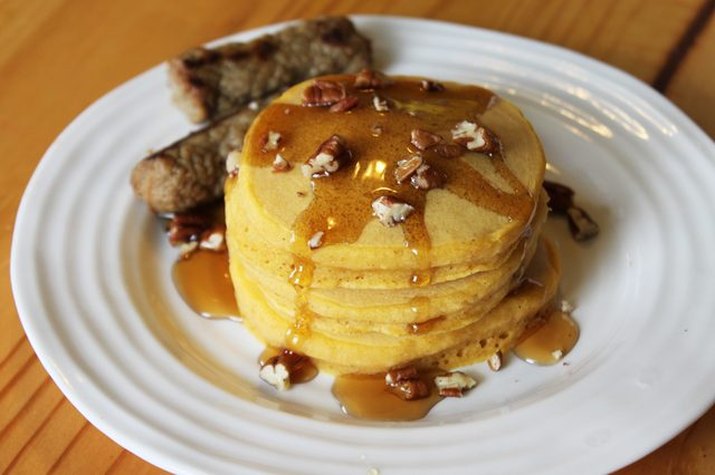 A stack of orange-hued pumpkin pancakes topped with pecans and maple syrup, served with two sausage links.