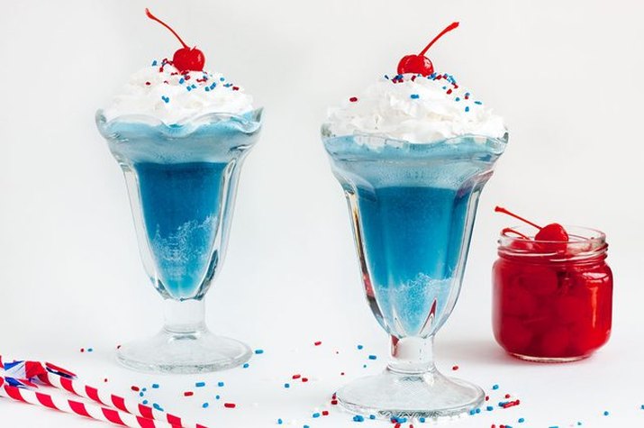 Ice cream soda glasses filled with blue-colored ginger ale, whipped cream and a cherry on top.