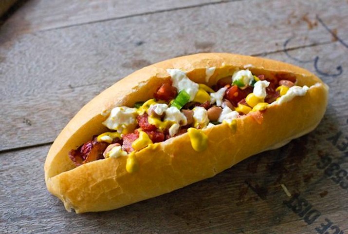 Sonoran hot dog wrapped in bacon and sprinkled with various toppings including pinto beans, grilled onions and tomatoes.